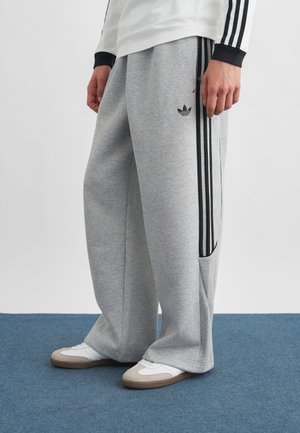 Person iført løse grå Adidas joggingbukser med sorte striber, hvide sneakers og en hvid langærmet top med sorte manchetter.