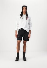 Hvid langærmet skjorte med farverige prints, sorte denim shorts med slidte detaljer, og sorte ankelstøvler. Casual streetwear stil.
