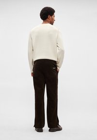 Krēmkrāsas džemperis ar apaļu izgriezumu, kombinēts ar tumši brūnām kordurojas biksēm ar sānu šķēlumiem un aizmugurējām kabatām. Melnas kurpes.