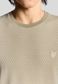 Beige Struktur-T-Shirt mit Rundhalsausschnitt und kleinem weißen Schmetterlingslogo auf der linken Brust. Weiches, gestricktes Material.