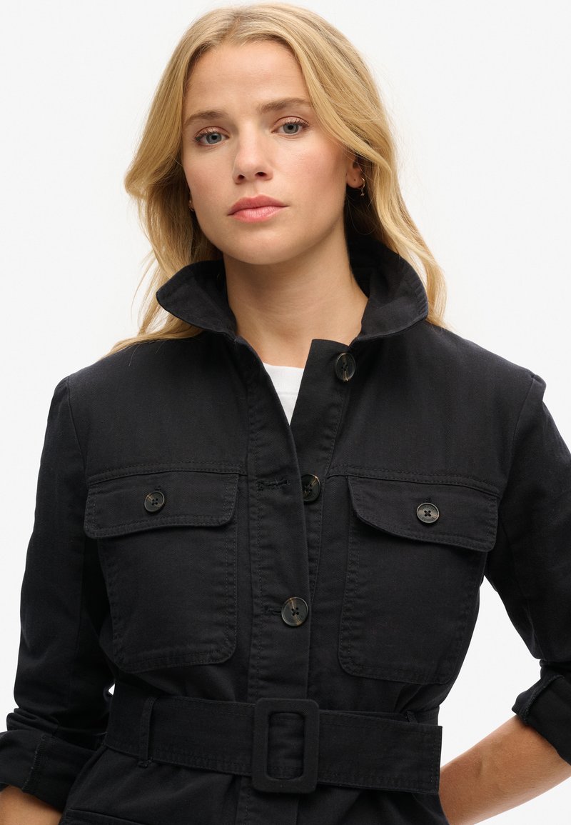 Chaqueta negra de estilo utilitario con cuello alto, mangas largas, dos bolsillos en el pecho y cintura ceñida con cinturón. Presenta cierre de botones y una tela texturada.