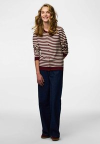 Svītraina bordo un krēma kardigans ar pogu priekšpusi, kopā ar vaļīgām tumšām džinsa biksēm un brūnām kurpēm. Modelis stāv neitrālā fona priekšā.