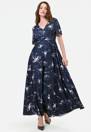 Donna che indossa un abito lungo blu navy con motivi floreali bianchi, maniche corte, scollo a V e tacchi marroni appuntiti.