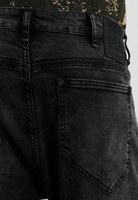 Pantalones vaqueros de mezclilla negra con un acabado texturizado, que presentan una etiqueta de cuero en la cinturilla y un diseño clásico de cinco bolsillos.