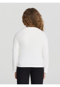 Biała bluzka z długim rękawem, okrągłym dekoltem i luźnym krojem. Miękki, strukturalny materiał z bocznymi rozcięciami u dołu. Noszona z czarnymi spodniami.