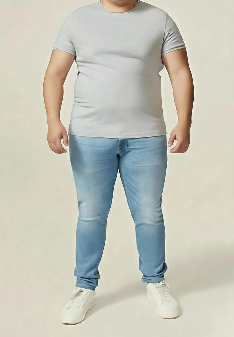 Erwachsener trägt graues eng anliegendes T-Shirt, hellblaue Jeans und weiße Turnschuhe und steht vor einem schlichten, neutralen Hintergrund.