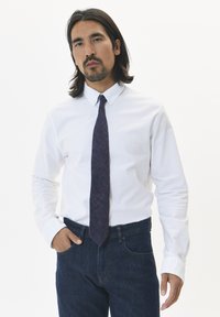 Chemise blanche à manches longues et boutons, associée à une cravate texturée violet foncé. L'individu se tient avec les mains dans les poches, portant un jean en denim foncé.