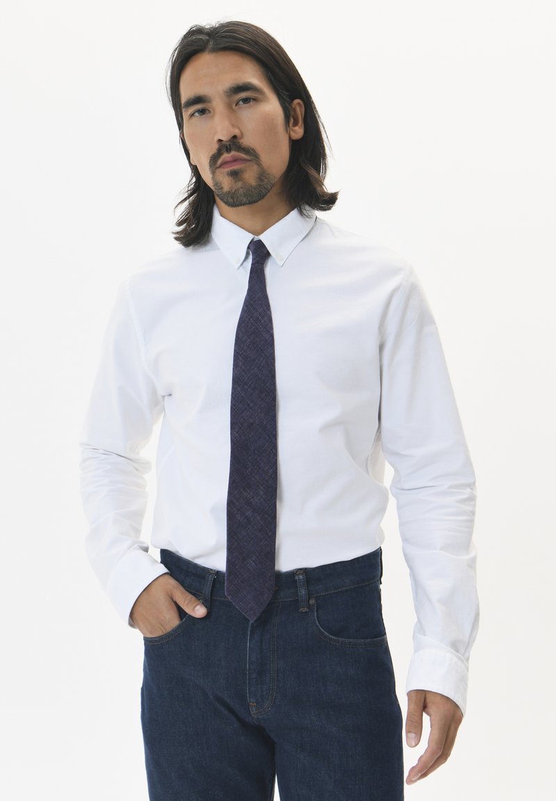Chemise blanche à manches longues et boutons, associée à une cravate texturée violet foncé. L'individu se tient avec les mains dans les poches, portant un jean en denim foncé.
