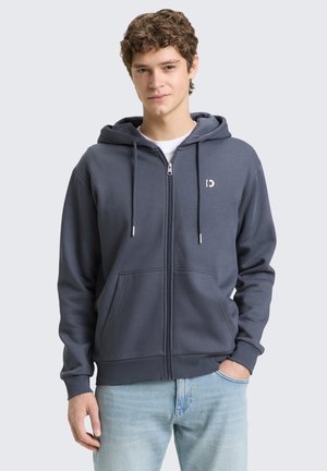 Junger Mann mit lockigem Haar, der einen dunkelgrauen Kapuzenpullover mit Reißverschluss und hellblaue Jeans trägt, steht mit einer Hand in der Tasche vor einem schlichten Hintergrund.