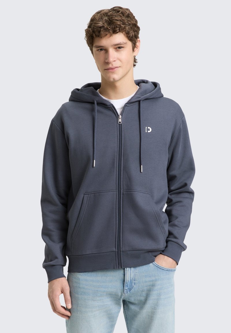 Jeune homme aux cheveux bouclés portant un sweat à capuche gris foncé zippé et un jean bleu clair, debout avec une main dans la poche, sur un fond uni.