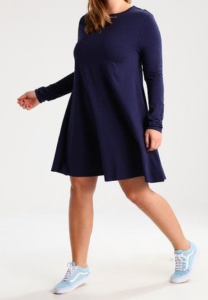 Donna che indossa un vestito blu navy a maniche lunghe e sneakers azzurre, in piedi con una gamba estesa in avanti contro uno sfondo uniforme.