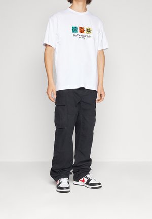 Uomo che indossa una T-shirt bianca con stampa grafica di icone solari colorate, pantaloni cargo neri e sneakers nere, bianche e rosse, in piedi su sfondo bianco.