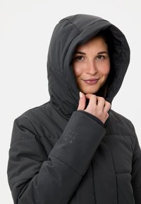Veste matelassée noire avec capuche, dotée d'un design matelassé et d'une texture lisse. Détail logo notable sur la manche et fermeture éclair visible.