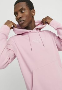 Pink hoodie made of soft fabric, featuring a front pocket, drawstring hood, and embroidered logo in white. Relaxed fit with ribbed cuffs.