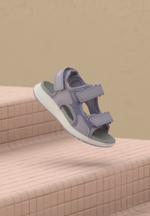 Sandale lilas clair à bout ouvert avec deux sangles ajustables, flottant au-dessus d'escaliers carrelés roses sur fond beige.