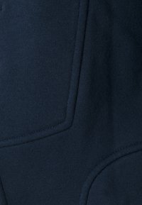 Gros plan sur un tissu bleu marine foncé avec couture et piqûre visibles, probablement une partie d'une poche sur un vêtement comme un pantalon ou une veste.