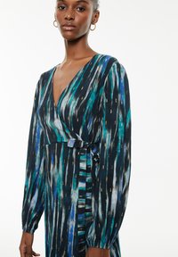 Vestido envolvente de manga larga en negro con patrones verticales en azul, verde azulado y dorado. Presenta un escote en V profundo y un lazo de tela en la cintura. Textura suave.
