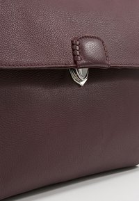 Gros plan d’un rabat de sac en cuir bordeaux texturé avec languette cousue et fermoir triangulaire argenté sur surface grise.