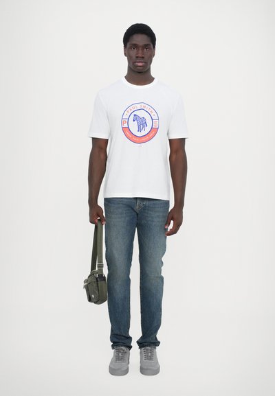 T-shirt en coton blanc avec un graphique circulaire représentant un zèbre et le texte "Paul Smith". Porté avec un jean bleu et un sac à épaule vert.