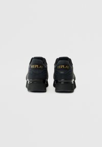 Zapatillas negras con un acabado de cuero suave, detalles de malla y una suela gruesa. Logo "REPLAY" en dorado en el talón.