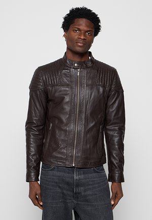 Mann trägt eine dunkelbraune Lederjacke mit Reißverschluss und gesteppte Schulterdetails sowie dunkelgraue Jeans, steht vor einem schlichten Hintergrund.