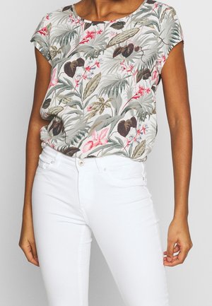 Femme portant un chemisier à manches courtes à motif floral dans des tons de vert, rose et marron, associé à un pantalon blanc taille haute.