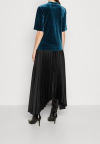 Tyrkysový sametový top s krátkým rukávem v kombinaci s černou skládací sukní, která se vyznačuje hladkou texturou a prodlouženou siluetou, nošený s lesklými černými botami na podpatku.