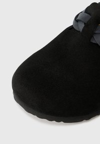 Black suede shoe with a rounded toe, featuring a woven gray accent on the top. The sole is textured and slightly raised.