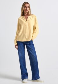 Blusa gialla a maniche lunghe con scollo a V e dettagli con bottoni, abbinata a jeans blu in denim larghi e sneakers bianche. Texture morbida, stile casual.