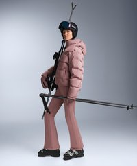 Žena u prašnjavo ružičastoj skijaškoj jakni i hlačama drži skije i štapove, nosi crnu skijašku kacigu, naočale, rukavice i crne skijaške čizme.