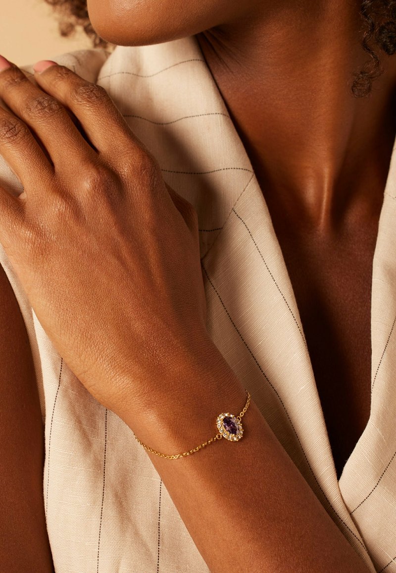 Gouden kettingarmband met een ovale paarse edelsteen omringd door kleine heldere stenen, gedragen om een zongebruinde pols.