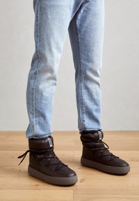 Schwarze gepolsterte High-Top-Sneaker mit grauen Gummisohlen, ausgestattet mit Schnürsenkeln und einem Etikett am Kn�öchel. Getragen mit hellblauen Jeans.