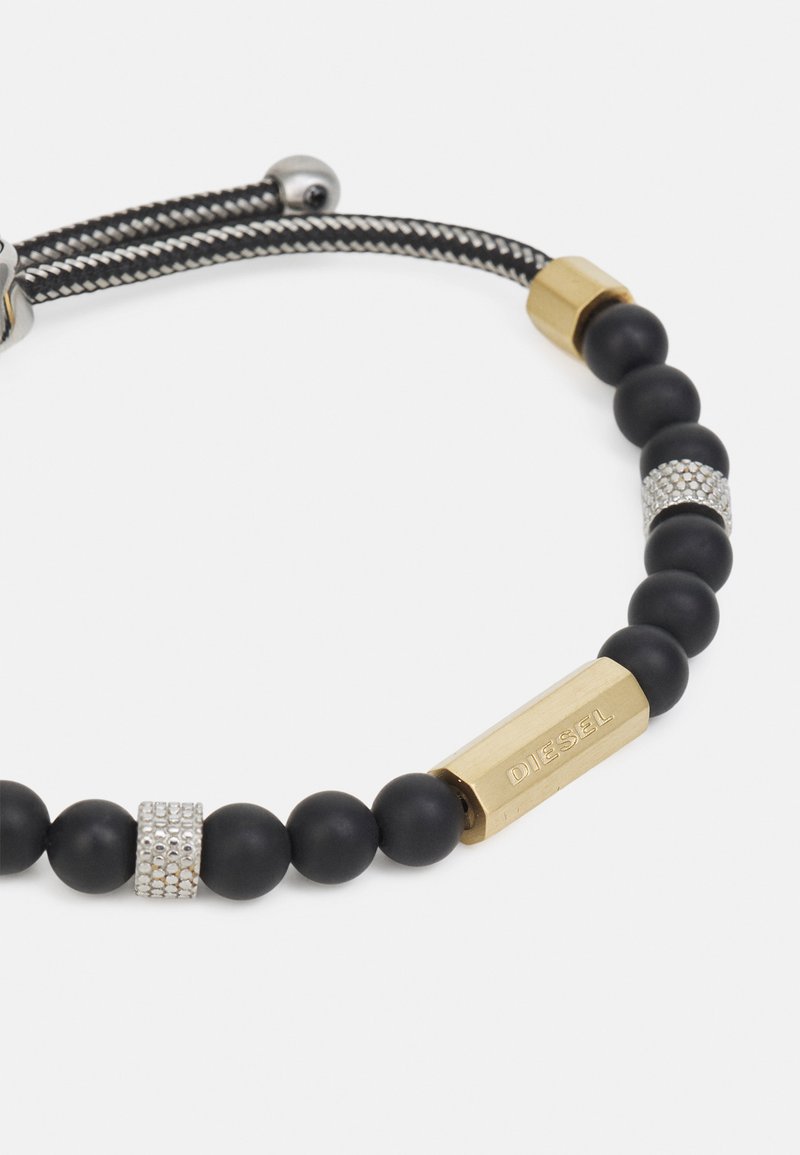 Schwarzes Perlenarmband mit Gummikugeln, Akzenten in Gold und Silber, versehen mit einem rechteckigen goldenen Anhänger, auf dem "DIESEL" eingraviert ist. Strukturierte Details vorhanden.