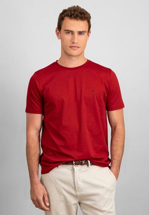 Red cotton t-shirt with a crew neck and short sleeves, featuring a small black logo on the left chest. Paired with beige trousers.