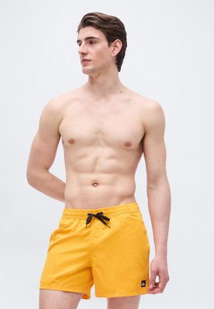 Nahý mladý muž s krátkymi hnedými vlasmi, oblečený v žltých plavkových šortkách s čiernym sťahovacím šnúrkou, smerujúci pohľadom do strany na jednoduchom pozadí.