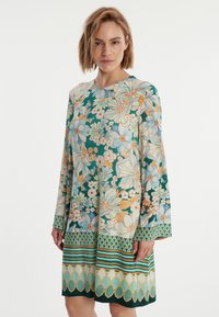 Robe fluide à fleurs en teal et crème avec de longues manches évasées, présentant un mélange de grands et petits motifs floraux, ainsi que des accents géométriques à l'ourlet.