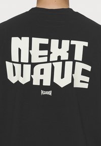 Czarna koszulka z bawełny z wyraźnymi, białymi literami w bloku "NEXT WAVE" na plecach, z logo "PEGADOR" poniżej.