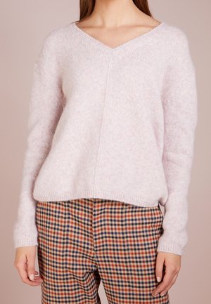 Personne portant un pull en peluche à col en V rose clair et un pantalon taille haute à carreaux beige, rouge et marine sur un fond neutre.