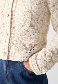 Texturierter Creme-Cardigan mit aufwendigen Häkelmustern, großen Knöpfen und einer lockeren Passform. Über Denim getragen, der das handgestrickte Design zur Geltung bringt.