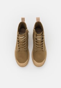 Botas de tobillo de lona marrón con suelas de goma beige. Presentan cordones, una cremallera lateral y una pestaña de tirón beige. Logo visible en la lengüeta.