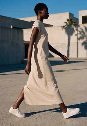 Une femme marche à l'extérieur en robe beige sans manches et baskets blanches devant des bâtiments en béton et un ciel bleu.