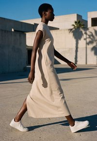 Woman walks outdoors in a beige sleeveless dress and white sneakers against concrete buildings and blue sky background.