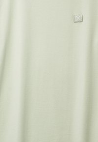 Vaaleanbeige kangas, jossa on pieni ommeltu neliskanttinen paikka, jossa on X-kuvio lähellä yläoikeaa kulmaa.