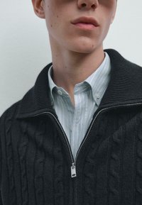 Tmavě šedý pletený svetr na zip s texturovaným kabelovým vzorem, nošený přes světle modrou pruhovanou košili s knoflíky.