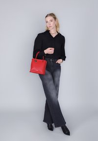 Rode leren handtas met twee handvatten, rechthoekige vorm, gestructureerd oppervlak. Model draagt een zwarte blouse en grijze flare jeans, met zwarte schoenen.