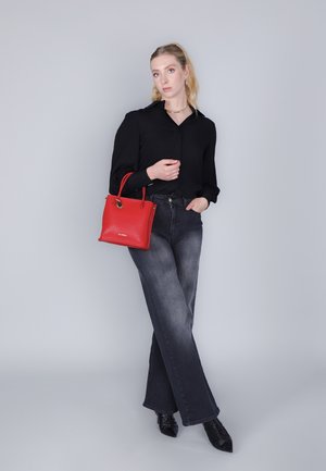 Borsa a mano in pelle rossa con due manici, forma rettangolare, superficie testurizzata. Il modello indossa una blusa nera e jeans svasati grigi, con scarpe nere.