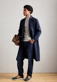 Homme portant un manteau long marin, un col roulé gris clair, un jean foncé, des baskets marron et noires, tenant un sac en cuir marron, debout sur un sol en bois.