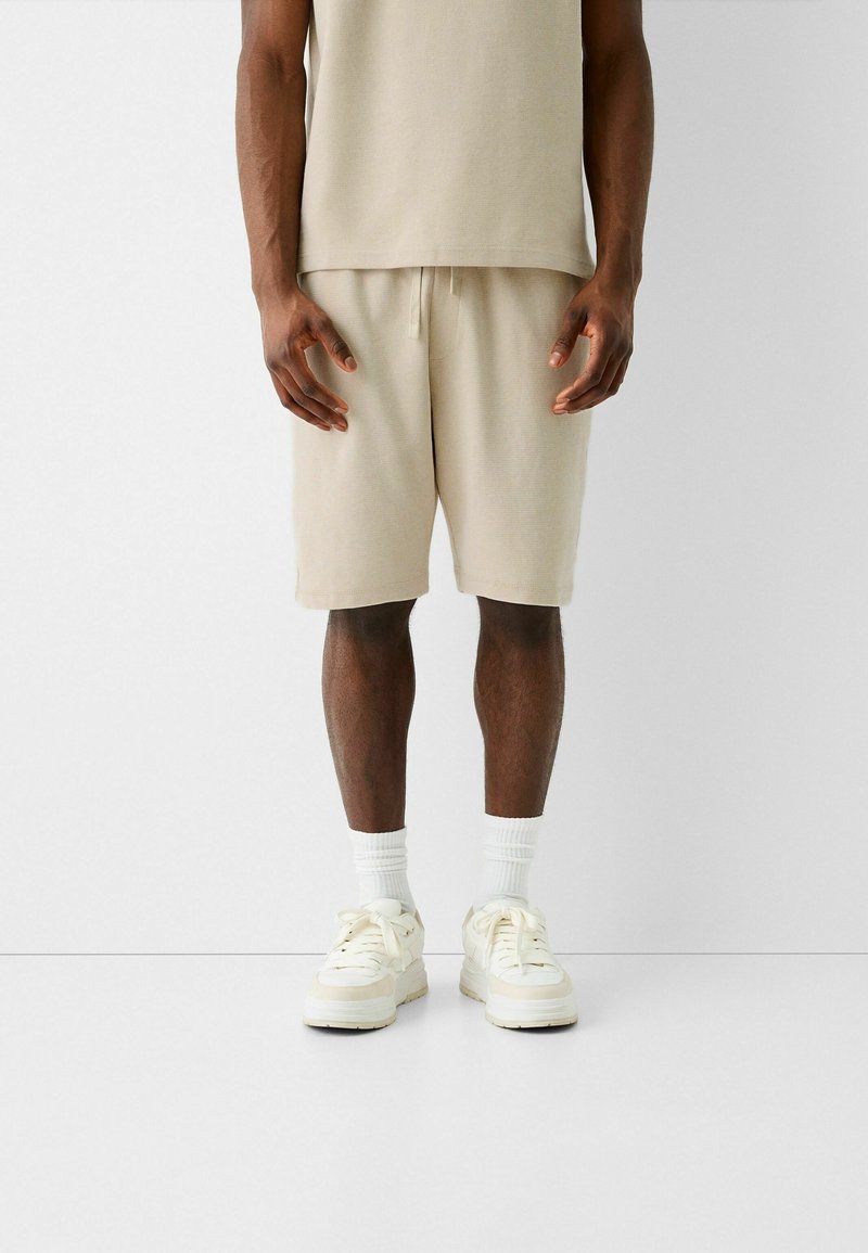 Short beige en coton avec une ceinture élastique, associé à un haut à manches courtes assorti. Des baskets blanches et des chaussettes côtelées complètent ce look.