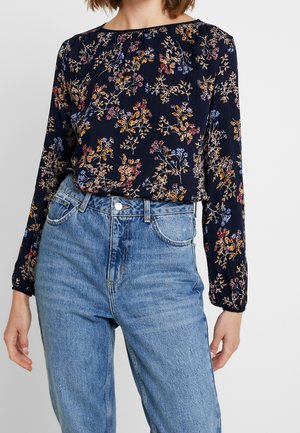 Mujer con una blusa floral de manga larga de color azul marino metida dentro de unos vaqueros de mezclilla de tiro alto de color azul claro, sobre un fondo liso.