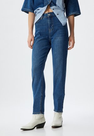 Højtaljede blå denimjeans med lige pasform, der har bæltestropper, forlommer og en glat tekstur. Båret med hvide ankelstøvler.
